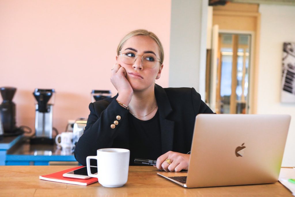 woman sitting at desk looking at laptop with her chin in her hand with a white coffee cup on desk