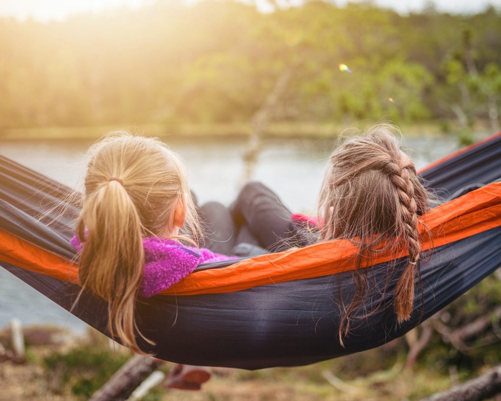 two young girls in a hammock next to a lake with sunshine and trees in background