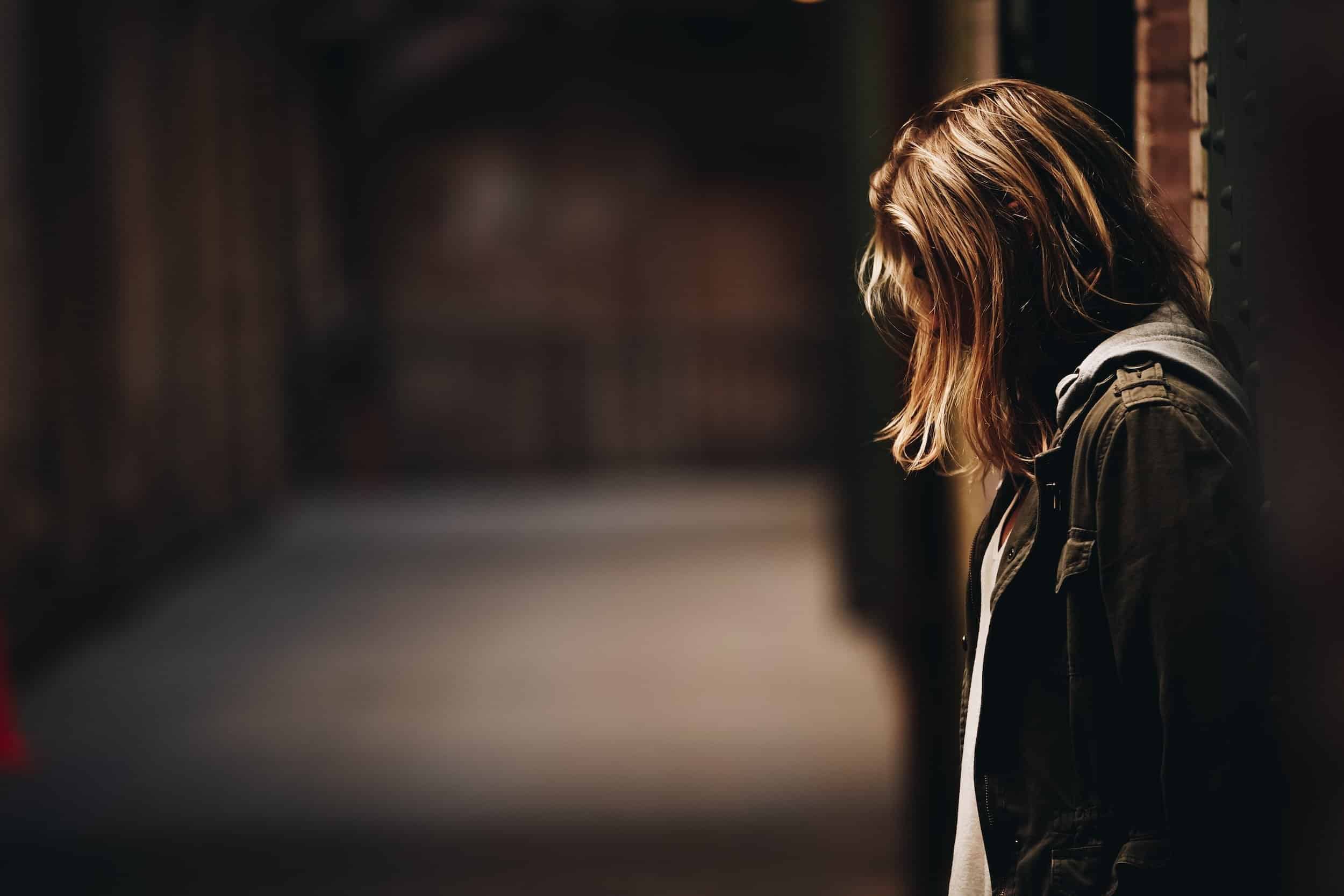 young woman leaning against wall in low lighting, with head bent down