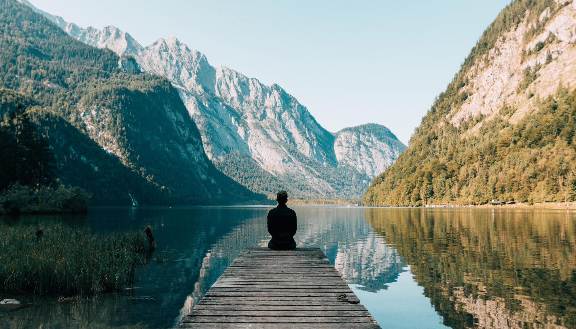 man sitting at end of dock on lake looking at mountains with snow