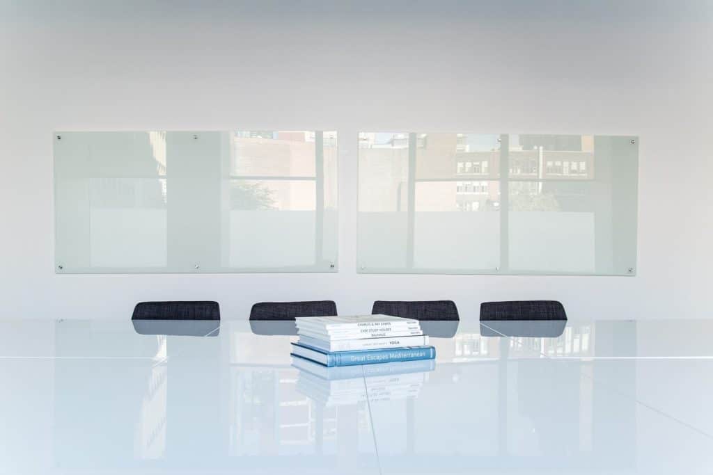 glass desktop with books resting on it with glass windows in background and four black chairs