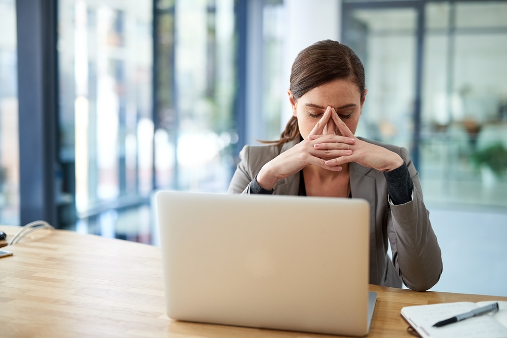 woman at desk in front of laptop with eyes closed leaning into clasped hands